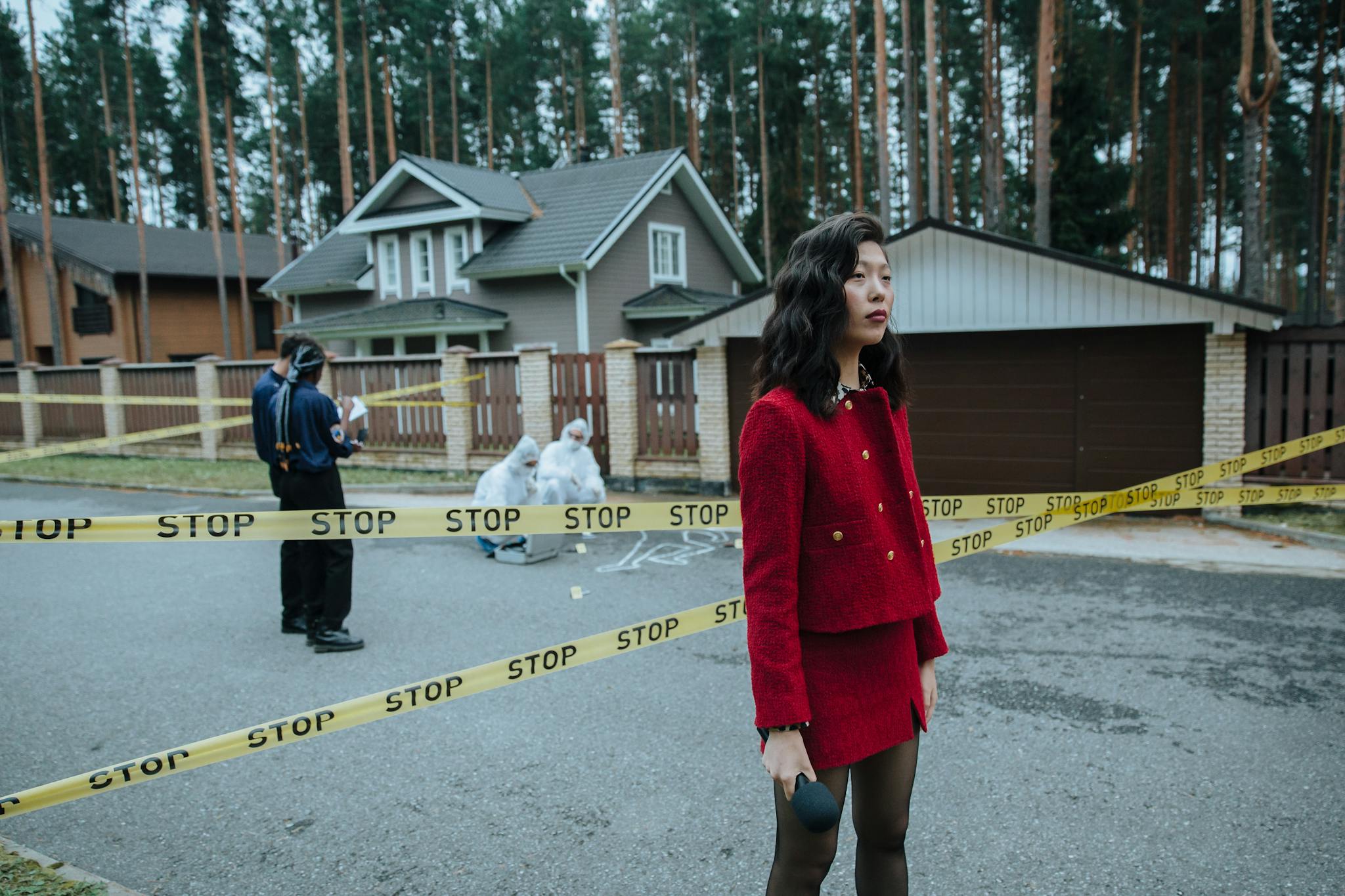 Our Mission A woman reporter in a red outfit at a crime scene marked with caution tape, outdoors near houses.